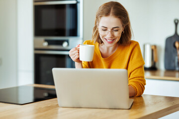 cheerful young woman uses a laptop at home in kitchen with  cup of morning coffee
