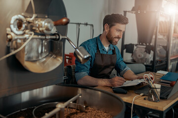 Handsome barista in uniform working with documents on small coffee factory 