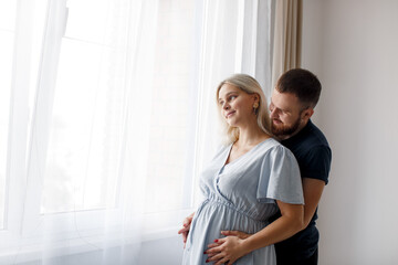 Fototapeta premium A man hugs his pregnant wife from behind and touches his belly near the window of the house. Pregnant couple. Lifestyle. 