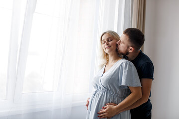 Naklejka premium A man hugs and kisses his pregnant wife from behind and touches his belly near the window of the house. Pregnant couple. Lifestyle. 