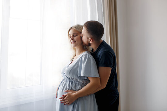 A man hugs and kisses his pregnant wife from behind and touches his belly near the window of the house. Pregnant couple. Lifestyle. 