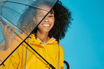 Portrait of smiling African woman with afro-hairstyle in yellow raincoat isolated over blue...