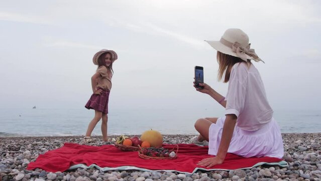 Woman taking pictures of her little pretty daughter by the sea on her phone