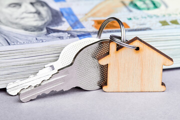 Trinket in the form of a wooden house with the keys to the apartment on the background of a stack of one hundred dollar bills