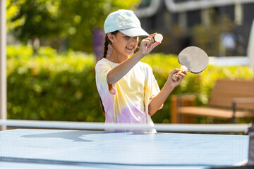 Young teenager girl playing ping pong. She holds a ball and a racket in her hands. Playing table tennis outdoors in the yard. - obrazy, fototapety, plakaty