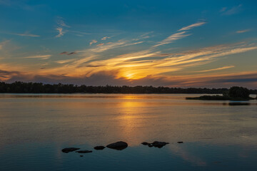 Beautiful sunset over the Dnieper River. Yellow cirrus clouds over the forest. Sun light reflection and stones in water