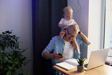 Young father working remotely on laptop with little baby son in his arms