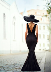 Fashion Woman in Black Hat and Evening Dress Back side view. Elegant Lady in Long Gown Rear view. Old Fashioned Model Looking away at City Background. Mysterious Women Portrait
