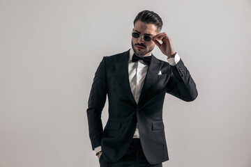 confident groom in elegant tuxedo with hand in pocket adjusting sunglasses