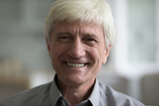 Happy handsome older grey haired Caucasian man posing indoors, looking at camera with toothy cheerful smile. Close up of positive male face with wrinkles. Indoor headshot portrait