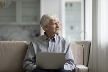 Thoughtful focused senior retired man holding laptop computer, looking away in deep thoughts, thinking over problem, trouble, sitting on sofa at home, feeling worried, concerned. Communication