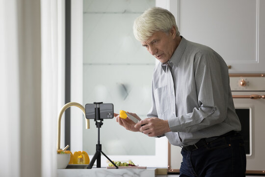 Serious mature older man filming cooking dinner for blog, watching video recipe of salad, recording workshop, showing slice of food at smartphone camera, presenting organic ingredients