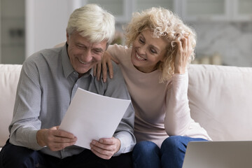 Cheerful old mature married couple reading paper document at laptop on home couch, reviewing...