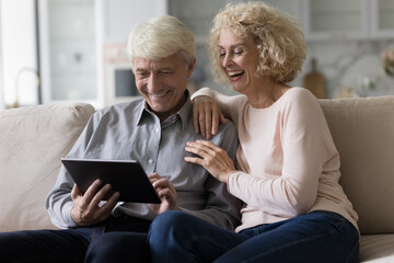 Cheerful mature retired couple shopping on Internet with ecommerce online app on tablet together, watching content, smiling, laughing, enjoying leisure with digital device, wireless communication