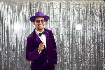 Happy funny man in purple velvet party suit, bow tie, hat and extravagant sunglasses smiles and...
