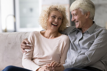 Fototapeta na wymiar Joyful mature old married couple enjoying leisure, dating at home, sitting on sofa, hugging with love, tenderness, affection, talking, laughing. Relationship, retirement