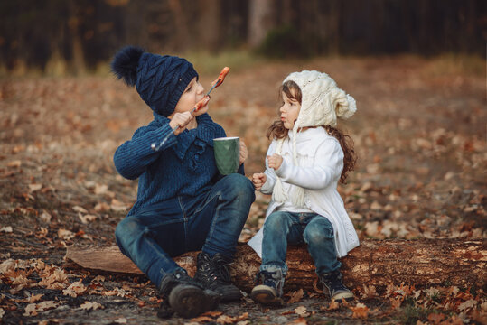 children in the autumn forest on a picnic, a boy and a girl, brother and sister sit on a log and eat sausages fried on a fire, drink cocoa