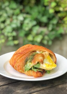 A Delicious Egg And Pesto Croissant Sandwich On A White Plate With Green Ivy In The Background