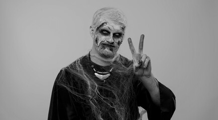Frightening man with Halloween zombie bloody wounded makeup showing victory sign, hoping for success and win, doing peace gesture, smiling with kind optimistic expression on studio gray background