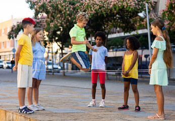 Multiracial kids playing together outdoors, jumping over rope. - obrazy, fototapety, plakaty