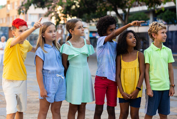 Group of multiracial kids standing outdoors during summer day, pointing fingers. Curious interested children gesturing.
