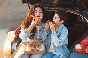 Funny mom with teen son sitting in the trunk of a car and eating slices of take away pizza from...
