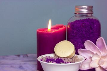Obraz na płótnie Canvas Violet lavender sea salt for bath in the bottle, a burning candle and branch of cotton flower. A bar of soap or dry shampoo. Face and body care, spa treatments concept. Cosmetic products. Copy space.