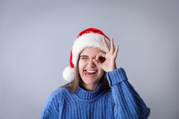 Playful woman in Santa hat and blue sweater showing OK gesture on grey background