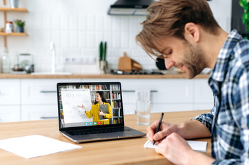 Online learning. Side view of a smart positive caucasian student guy sit at home in a kitchen with laptop, listening to an online lecture, on a screen caucasian girl teacher teaches a student remotely