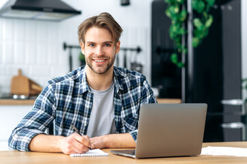 Successful clever caucasian young man, student, freelancer, programmer, sitting at home in kitchen, taking notes in a notebook during an online webinar or lecture, working on a project, using laptop