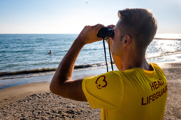 Lifeguard on the beach looking through binoculars. Safety while swimming, handsome brunette male...