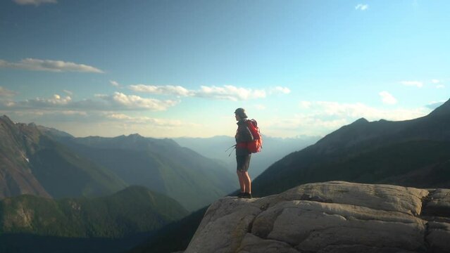 Caucasian man on hiking trip walks out to edge of cliff to view epic mountain landscape, wide