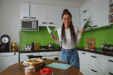 Joyful smiling mixed-race woman standing in kitchen holding knife and onion preparing food