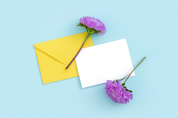 White blank card, yellow envelope and purple flower on blue background. Minimal style. Top view Flat lay Mockup