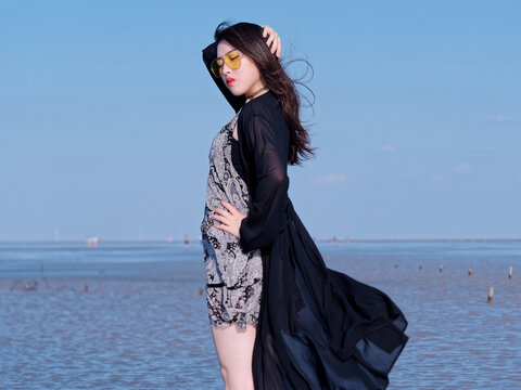 Portrait of beautiful young Chinese woman with black long hair in black dress posing with blue sky and beach background in sunny summer day. Carefree girl enjoy her free time. Beauty lifestyle.