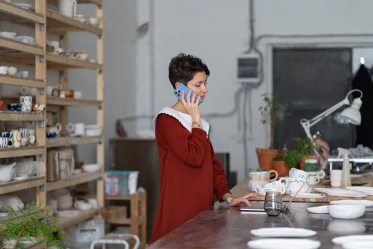 Young woman ceramics studio manager talking by phone and using laptop, female entrepreneur pottery shop owner ordering supplies and equipment, ceramic artist making phone call, standing in workshop