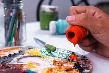 High quality photography. Paint pots of different colors, paint palette with a lot of stains and brushes in a glass jar. An artist's hand placing paint on an orange palette.
