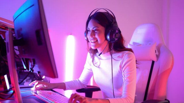 Happy pretty long-haired Asian transgender professional gamer wearing a gaming headset having fun playing online video games on a personal computer in room and raising fist up after winning the game.