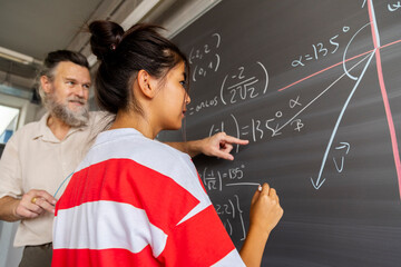Mature caucasian male high school maths teacher explains blackboard exercise to asian girl teen student. - obrazy, fototapety, plakaty