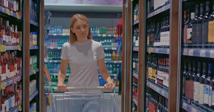 Beautiful girl in the department of alcoholic beverages chooses a bottle with a drink. Racks of alcoholic beverages stand in a row in the supermarket. 4k, ProRes