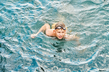 Fototapeta na wymiar Junior schoolboy in swimming goggles enjoys holidays. Schoolkid swims in clear blue sea with smiling and excited expression on face closeup