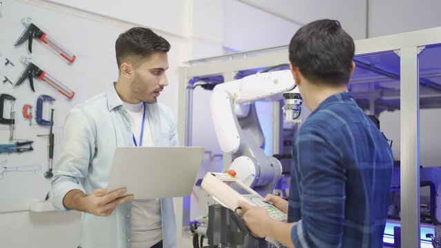 Young Asian male engineer showing the work of robotic arm machine with a robot controller to young Caucasian male apprentice engineer standing watching, asking, and recording details by laptop in hand