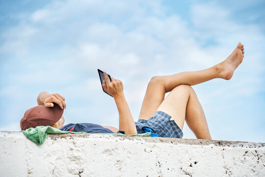 Young guy reads digital book on tablet lying on stone pier with bare legs against blue sky. Young lad enjoys reading uniting with nature at seaside