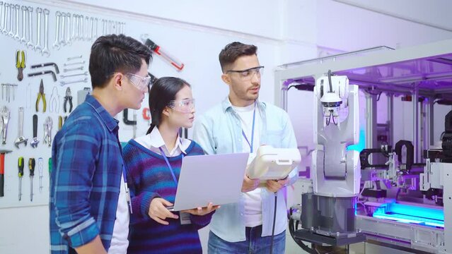Young Caucasian male engineer standing showing the work of robotic arm machine with a robot controller to young Asian male CEO and Asian female apprentice looking an information of machine in a laptop