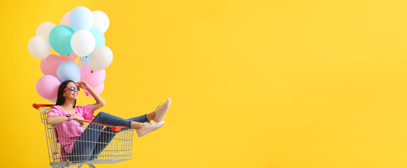 Fototapeta Portrait of fashionable young woman in shopping cart and with balloons on yellow background with space for text obraz
