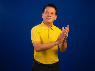 Portrait of smiling middle-aged asian man in casual yellow polo shirt applauding to camera isolated on blue background in studio, looking happiness and attractive professional leadership