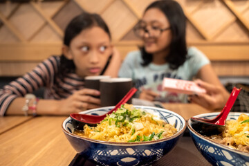 A bowl of noodle menu served at Japanese restaurant, with Asian teen girls having dinner together in the background