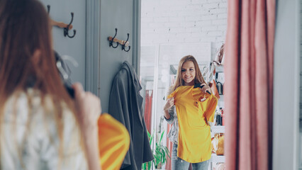 Mirror shot of young lady choosing clothes in fitting room. Girl is trying top checking size and length while standing opposite large mirror. Nice clothing store in background. - 528370130