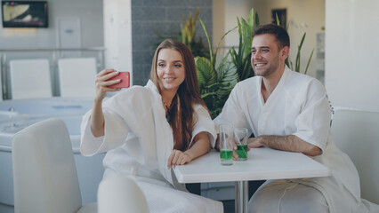 Obraz premium Pretty cheerful girl and her loving boyfriend are taking selfie with cocktail glasses using smartphone while relaxing in spa salon. They are smiling and posing looking at camera.