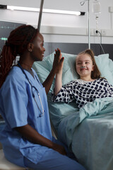Obraz premium Childcare healthcare facility staff high fiving ill kid under medical treatment. Nurse doing high five gesture with sick girl resting in hospital pediatric ward patient bed.